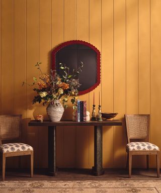 Warm and inviting hallway with mustard yellow tongue-and-groove paneling, a dark wood console table, and a red-framed mirror.