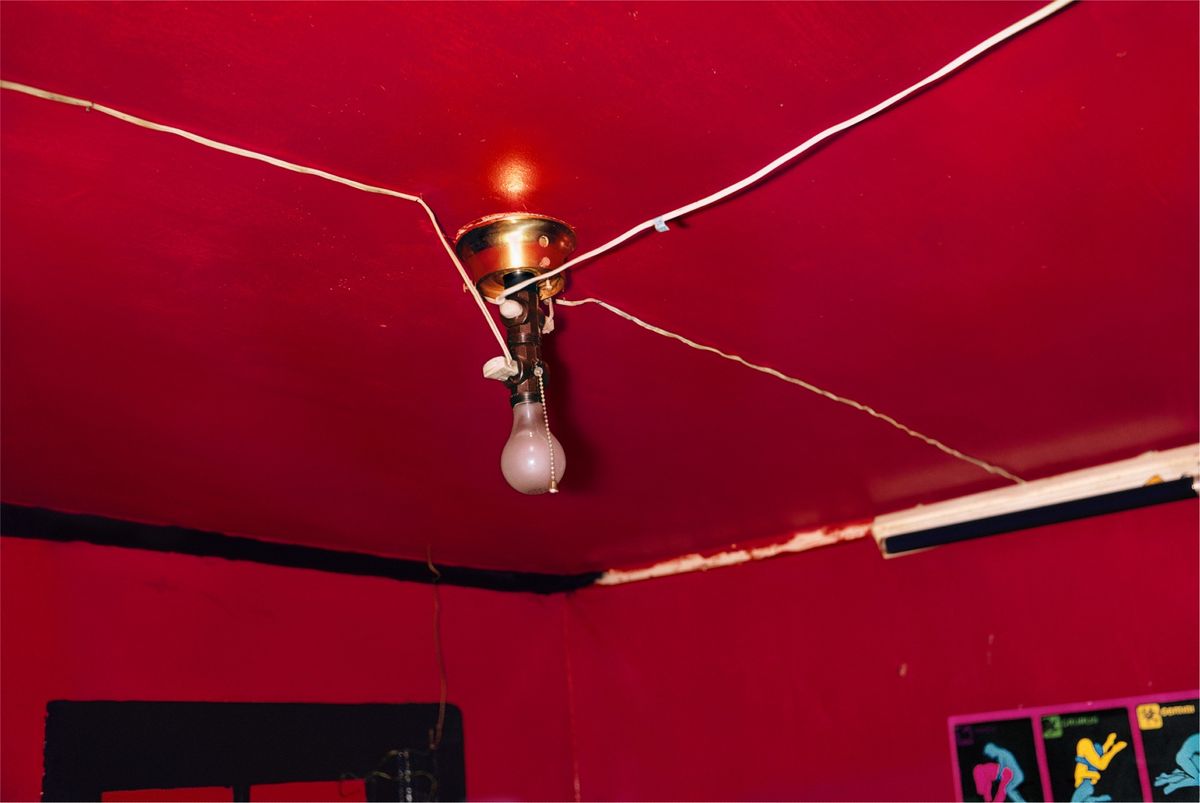 A color photograph of a bare lightbulb hanging on a cord from a deep red ceiling, with wires stretching across the room.