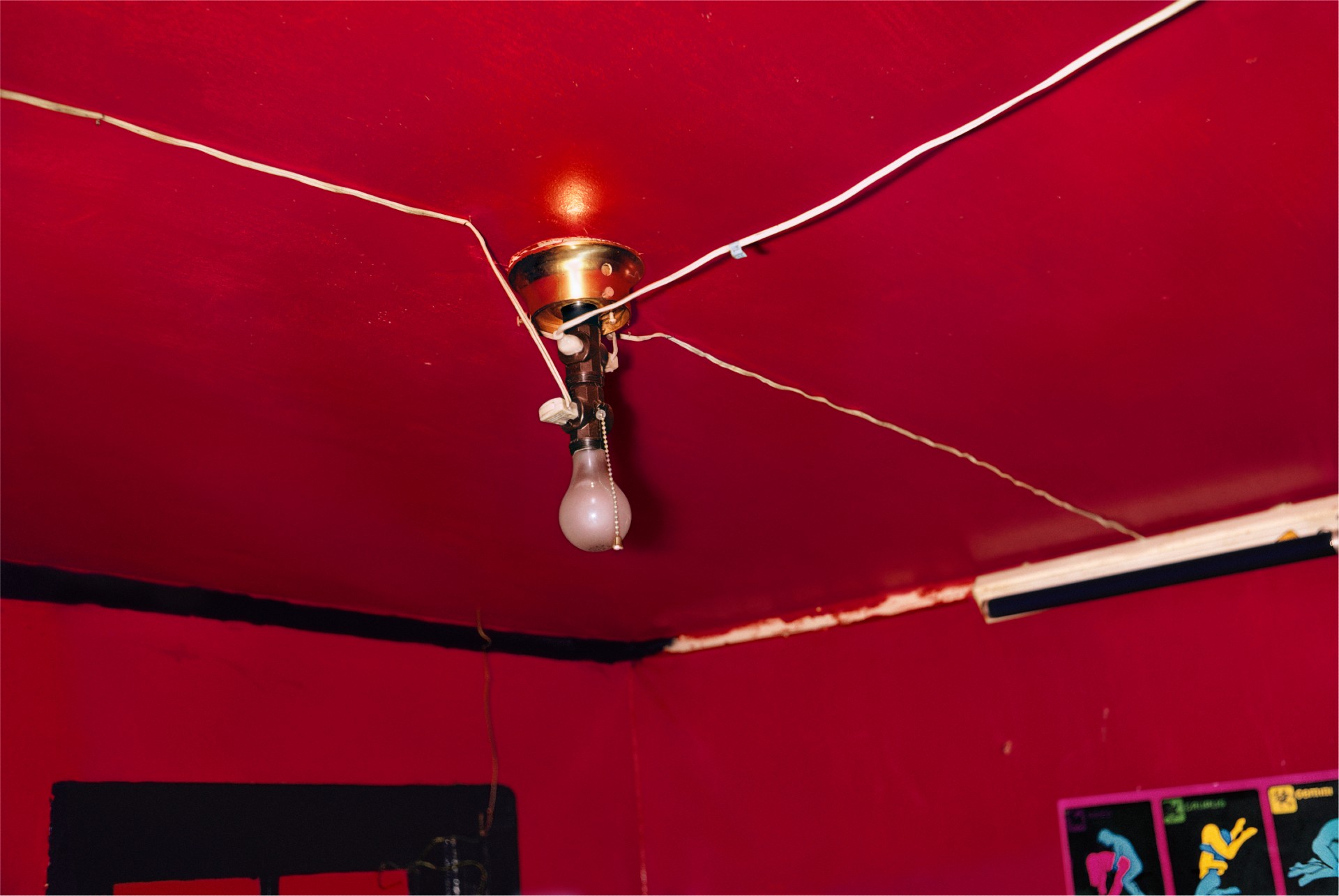 A color photograph of a bare lightbulb hanging on a cord from a deep red ceiling, with wires stretching across the room.