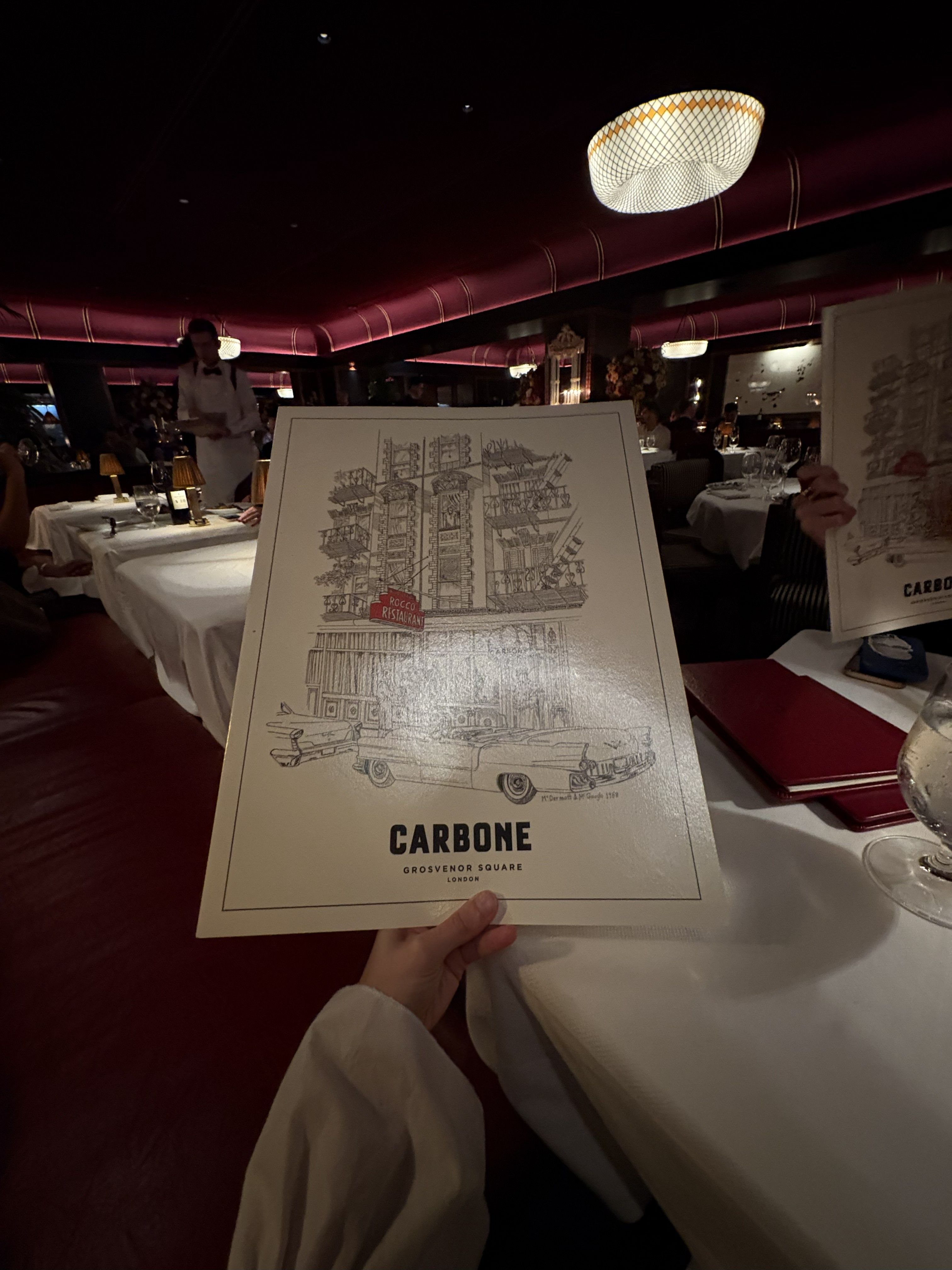Three image, the first depicting a hand as it holds a double-A3 restaurant menu with a handsketched restaurant exterior on its cover, the other depicting a cake trolley filled with desserts, and another capturing a hand holding a white dish topped with silver sauce boats.