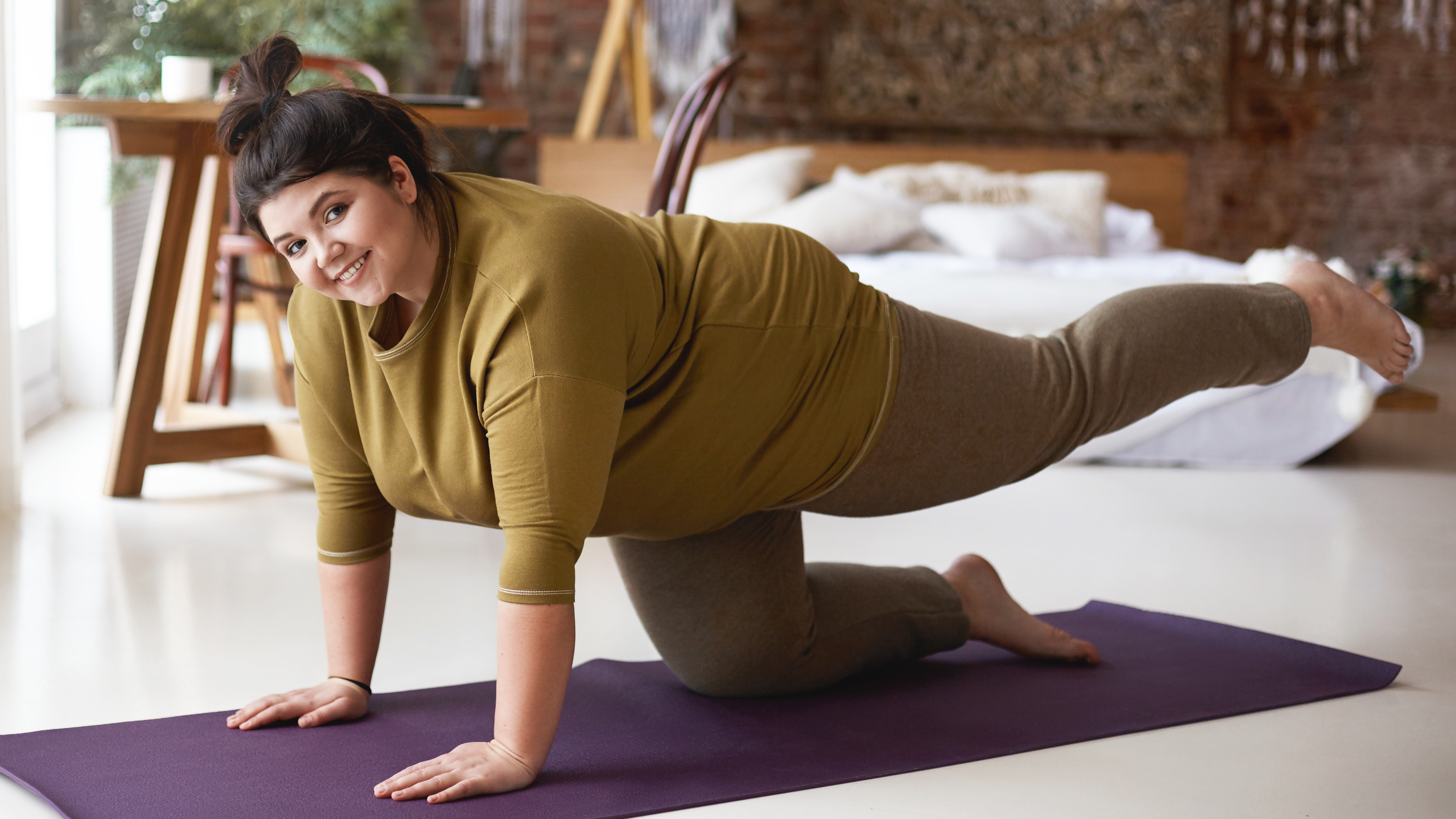 A woman smiles at the camera as she performs a four-point kneeling leg lift at home on a mat. Her hands and right knee are on the floor, while her left leg is extended behind her. 