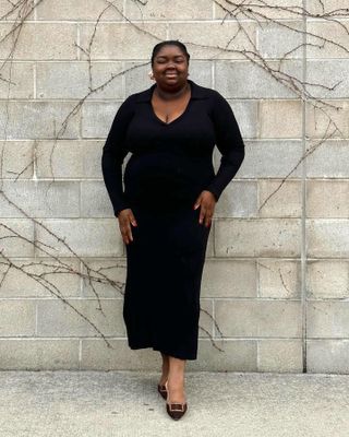 Fashion influencer ukuran plus Abisola Omole tersenyum sambil berpose dalam balutan gaun hitam lengan panjang dan sepatu hak mule Manolo.