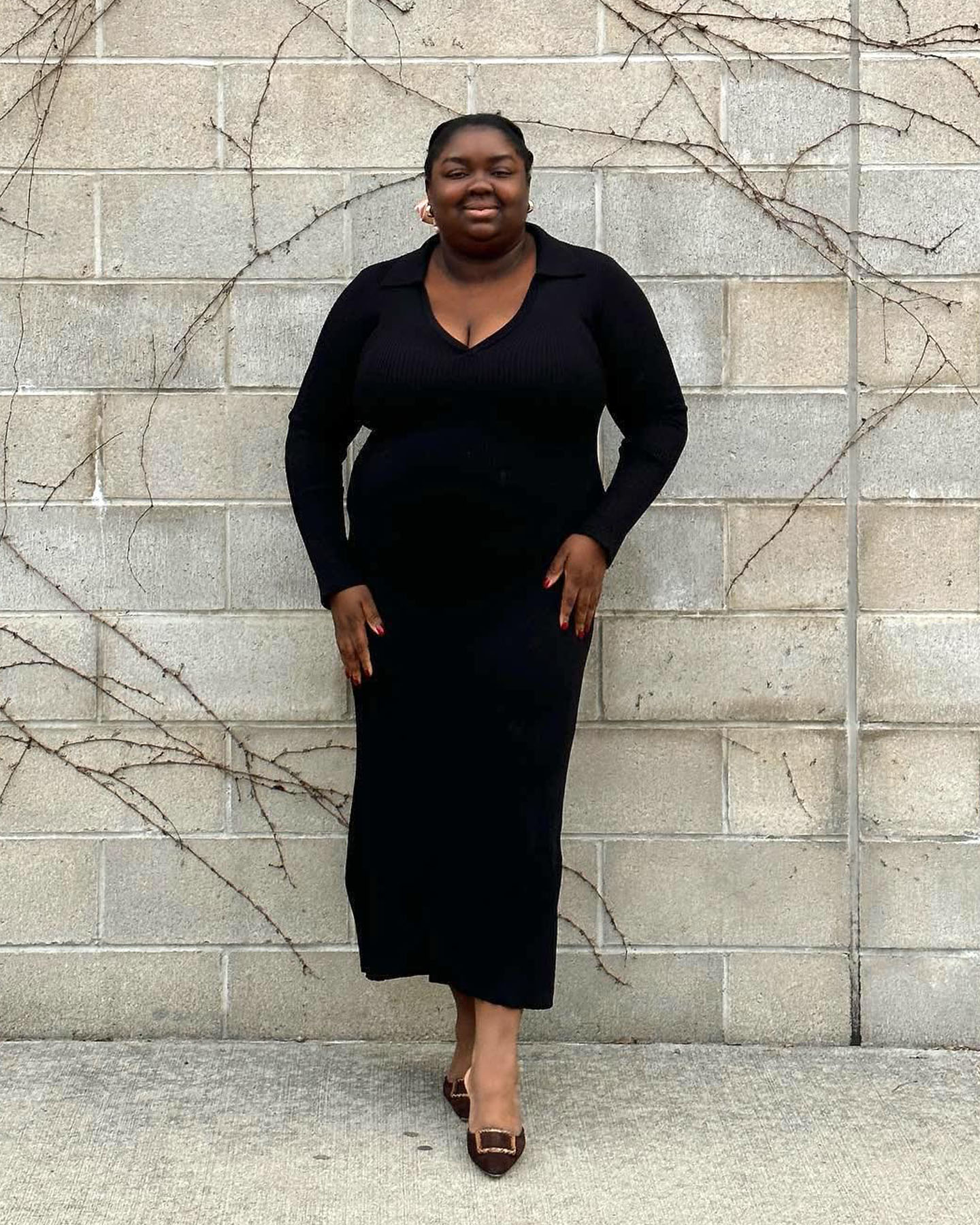 Plus size fashion influencer Abisola Omole smiles while posing in a long sleeve black dress and Manolo mule heels.