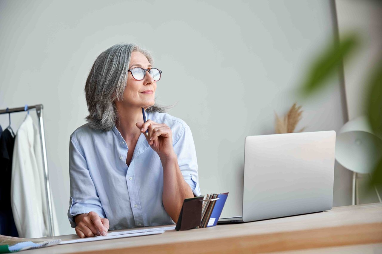 A woman sitting at a desk with a laptop wearing glasses and looking off in the distance.