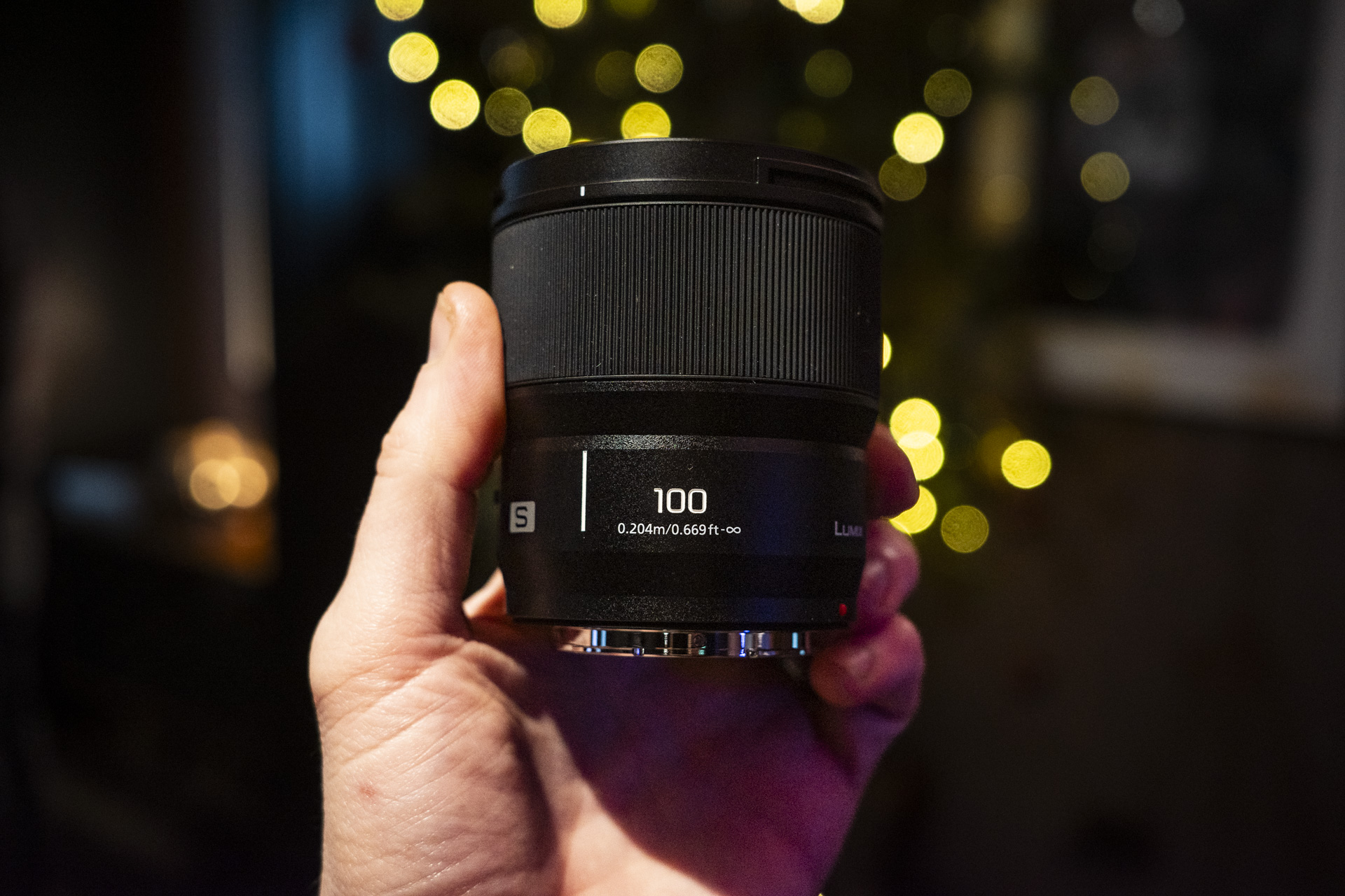 Panasonic Lumix S 100mm F2.8 Macro lens in the hand with Christmas lights background