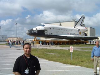SPACE.com Managing Editor Tariq Malik poses with the shuttle Atlantis in the background in July 2005 as it moved from a service hangar (far right in background) ahead of its STS-115 mission in Sept. 2006.