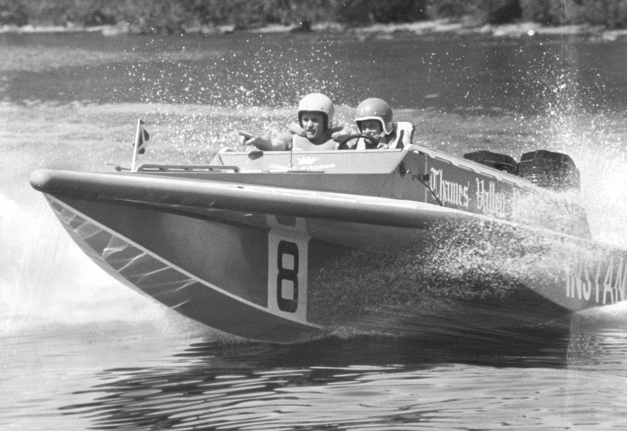 A black and white image of two people in helmets behind the wheel of a speedboat