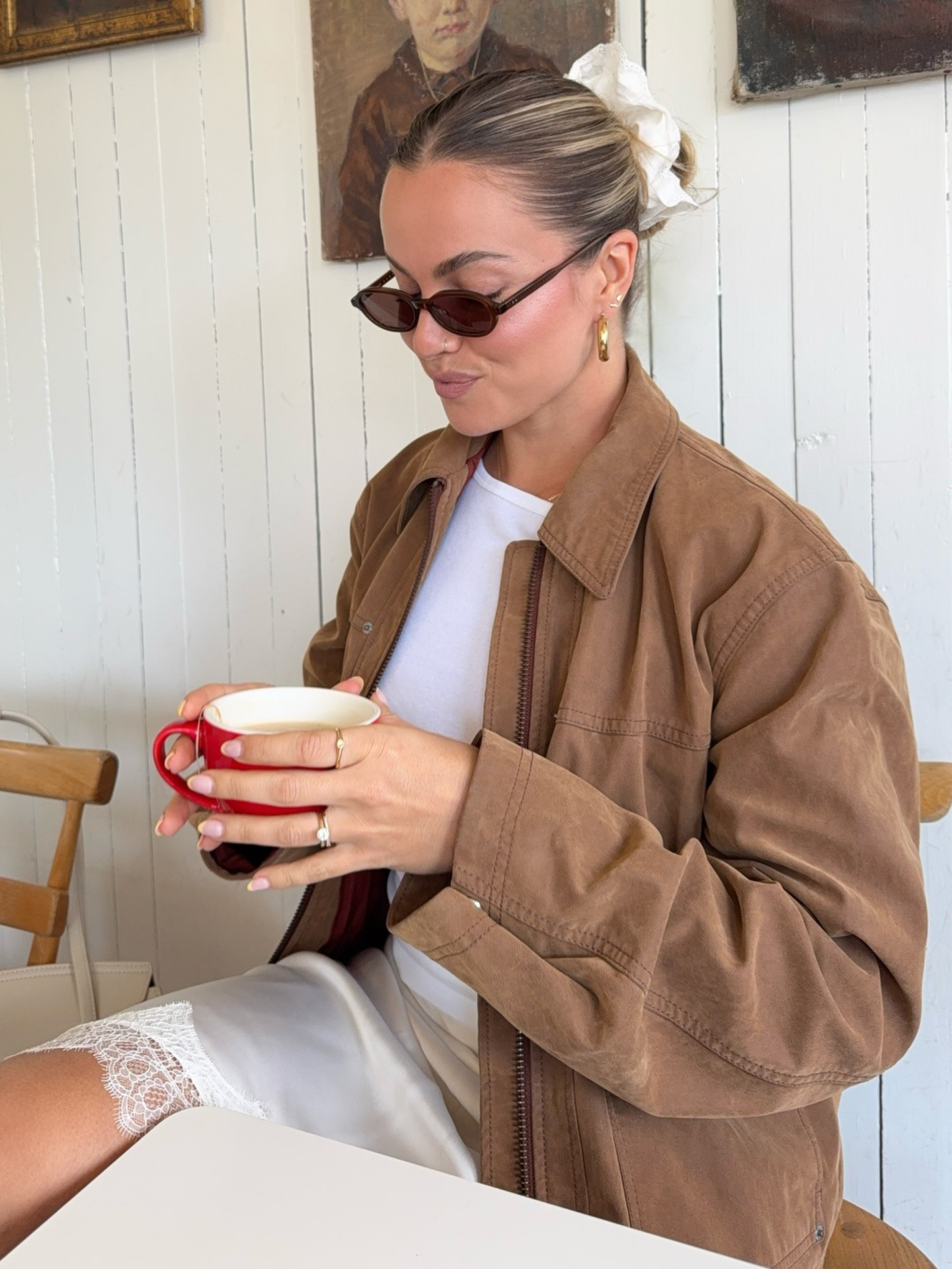 style influencer Izzy Dilger sits in a cafe holding a red mug wearing brown oval sunglasses, a white oversize scrunchie on her bun, a tan jacket, white T-shirt, and an ivory lace-trim slip skirt