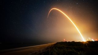 a time-lapse photo showing a streak from a rocket launch arcing over the beach at night.