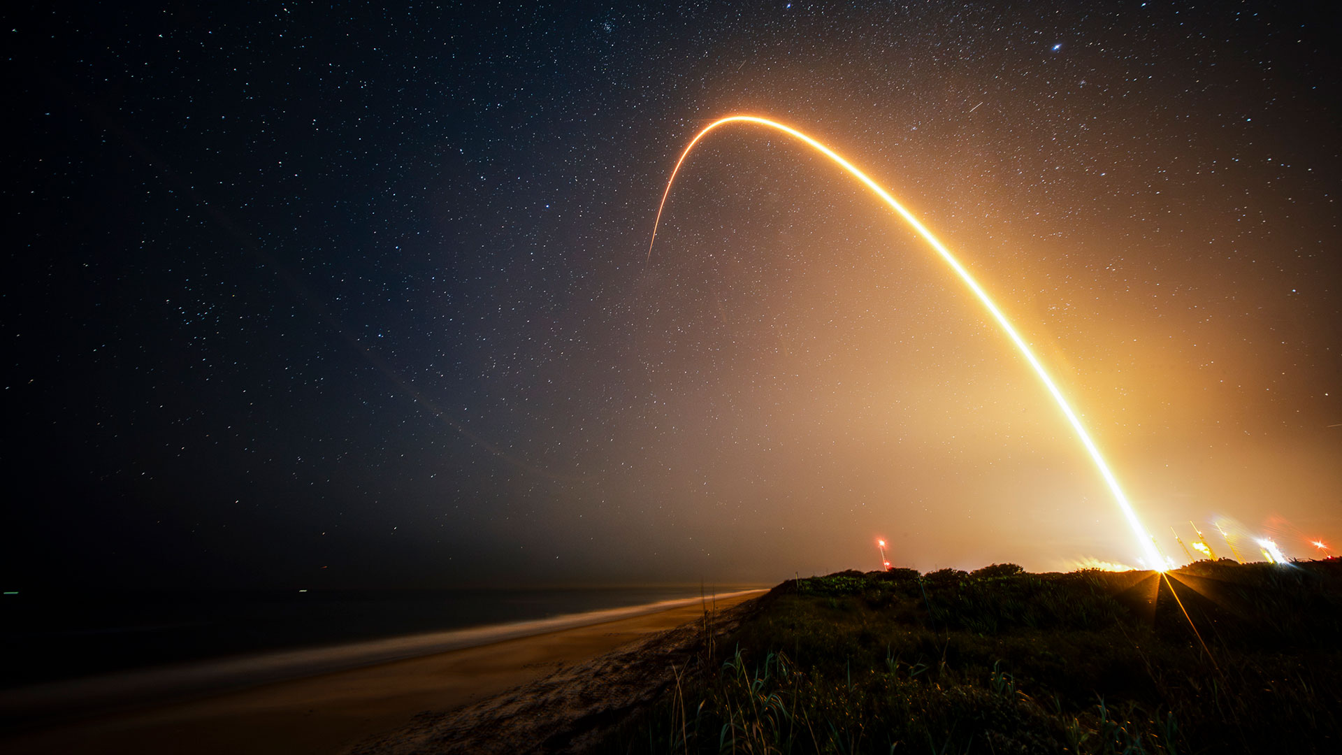 a time-lapse photo showing a streak from a rocket launch arcing over the beach at night.