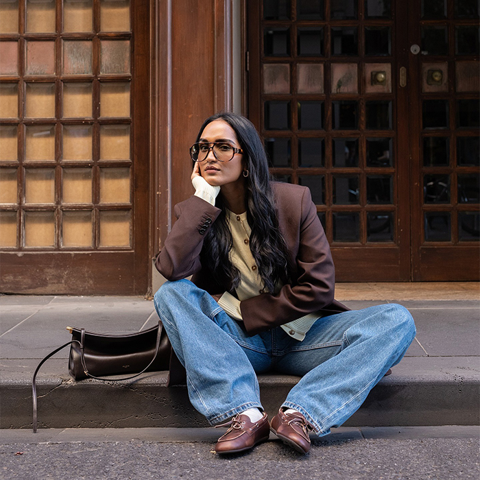 model wears brown blazer with cream cardigan blue jeans and brown loafers while sitting on a sidewalk and posing