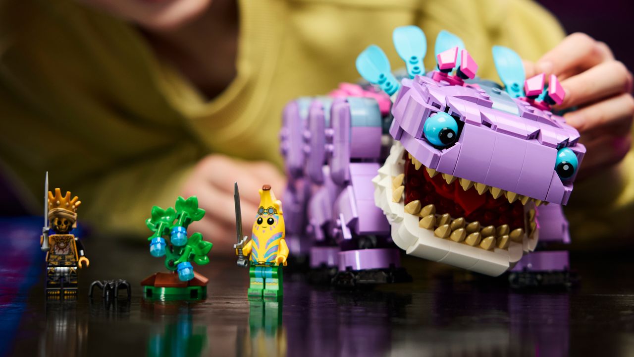 A hand adjusts the pose of Lego Klombo as it stands beside two minifigures