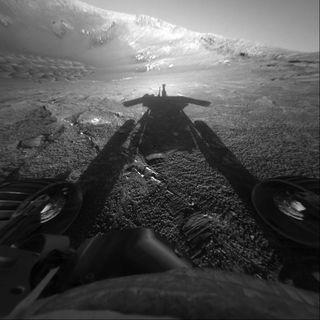 Opportunity rover on Mars looks away from the sun into Endurance Crater and sees its shadow. The image shows two wheels on the lower left and right, with the floor and walls of the unusual crater in the background. Although the companion Spirit rover has