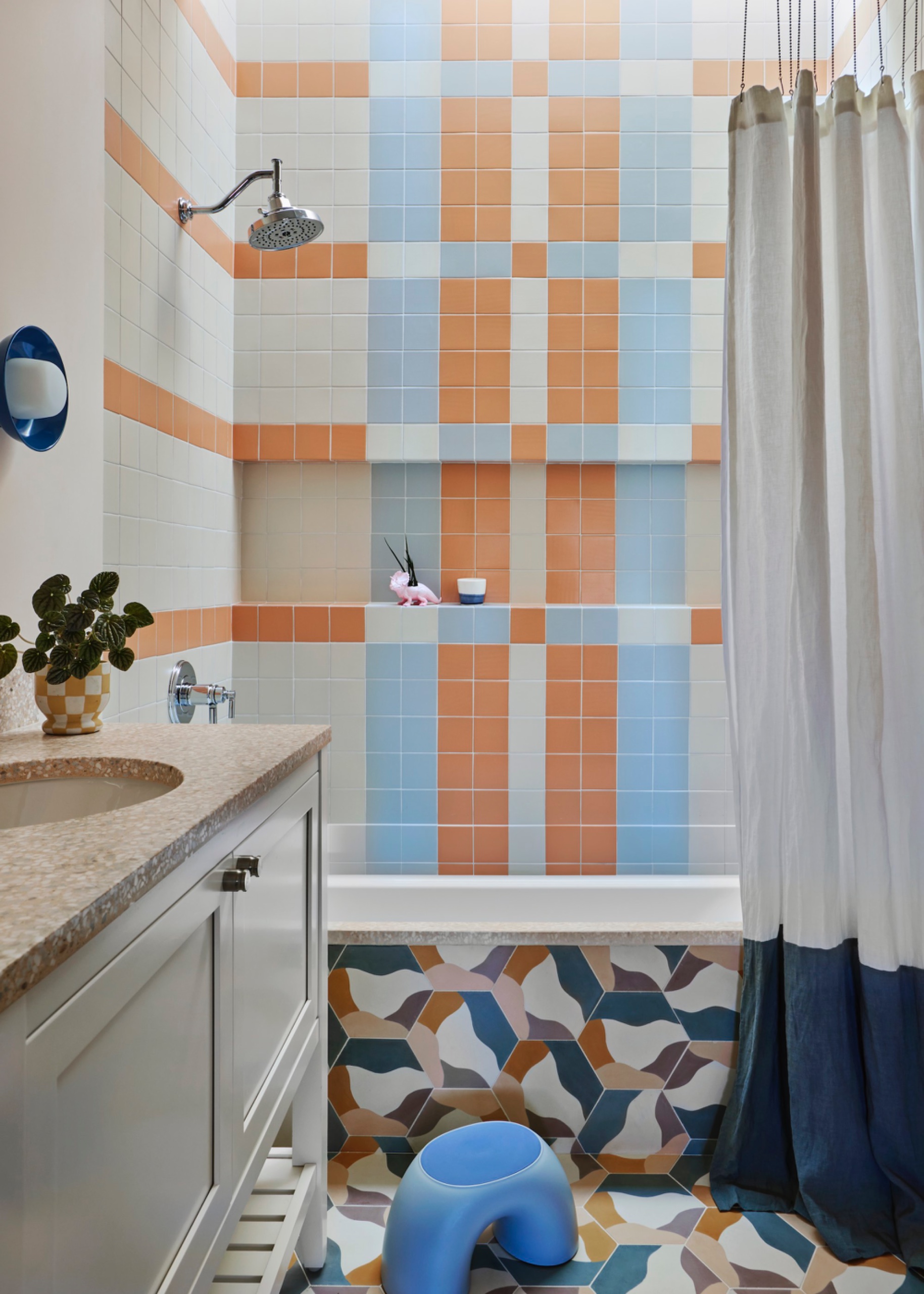Bathroom with orange, pale blue and cream square tiles arranged in plaid around a shower niche, patterned hexagonal floor and bath-front tiles, and a blue-and-white curtain.
