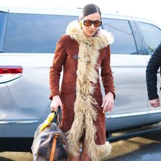 Bella Hadid wears a brown penny lane coat with chanel sunglasses and a louis vuitton duffle bag.