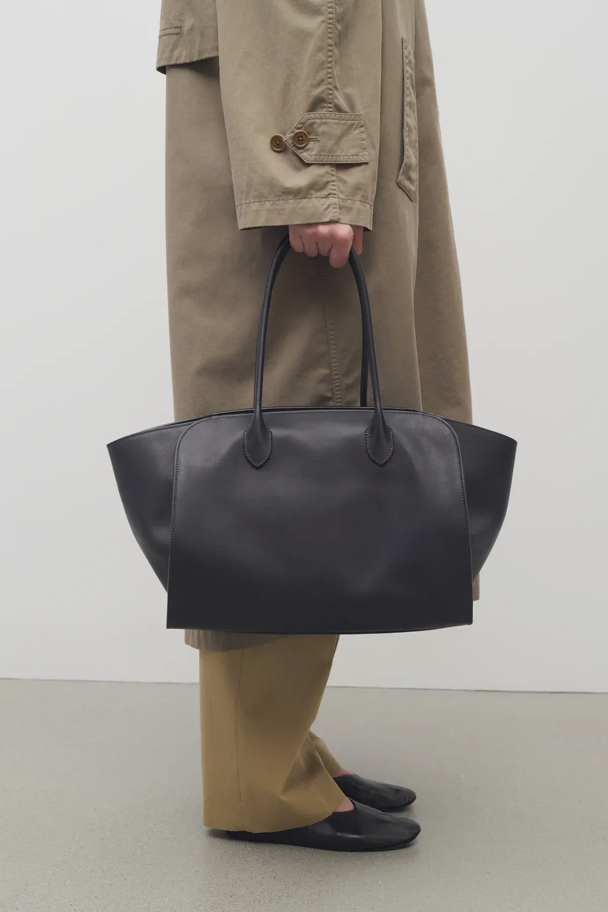 Person wears green trench coat, green trousers, black ballet flats and carries black bag