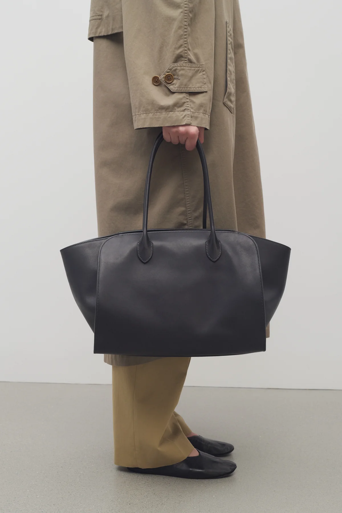 Person wears green trench coat, green trousers, black ballet flats and carries black bag
