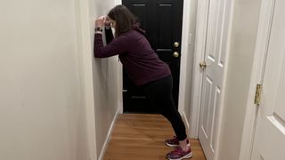 Trainer Jennifer Rizzuto performs a wall plank in an empty hallway. She leaning towards the wall, with her elbows bent and forearms resting on it. The rest of her body is held in a straight line, from her shoulders to her feet.