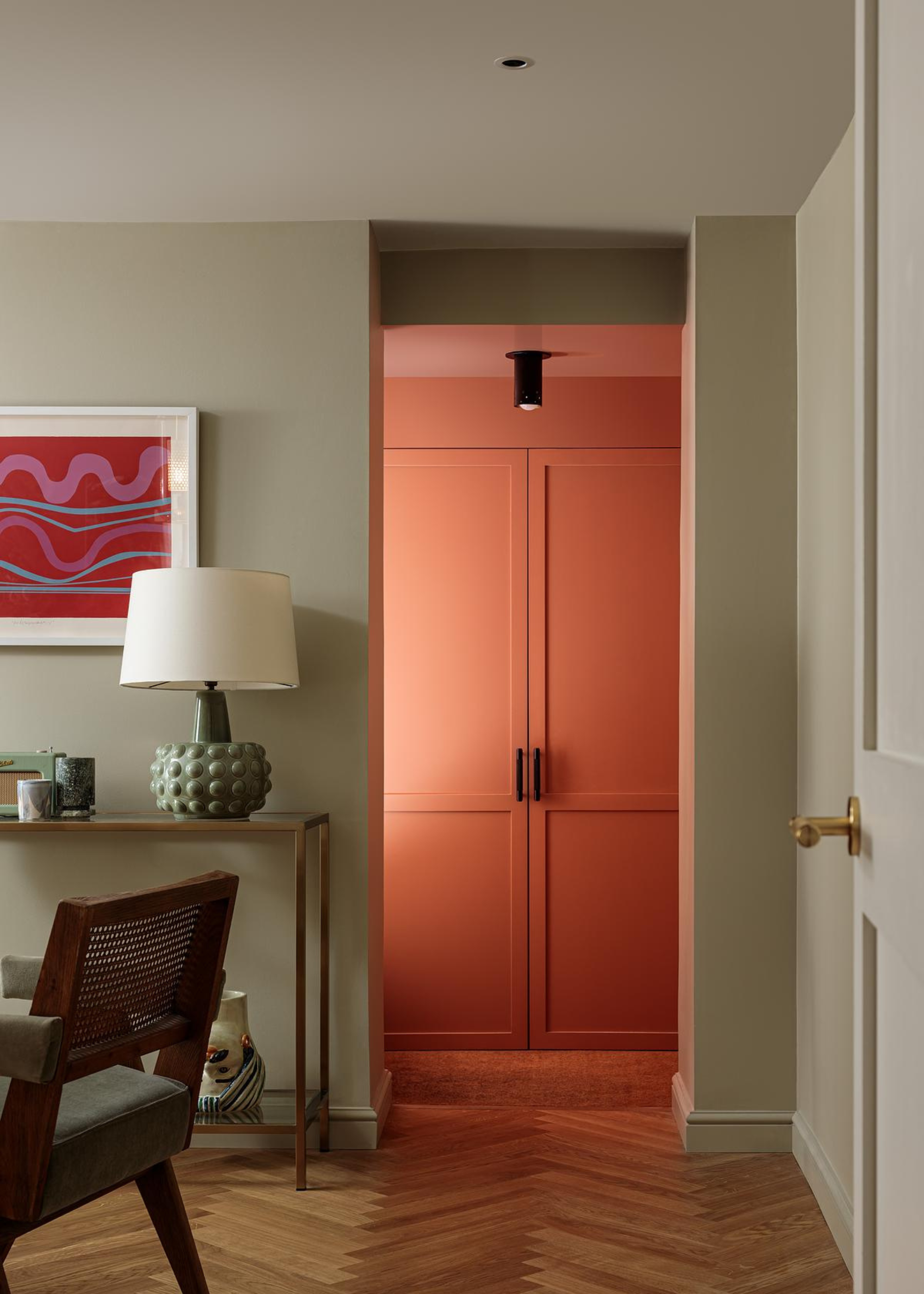 A living room leading to an orange cabinet with a focused overhead light. The room also holds a side table with a textured green lamp, a print on the sage green wall, and a wooden upholstered chair