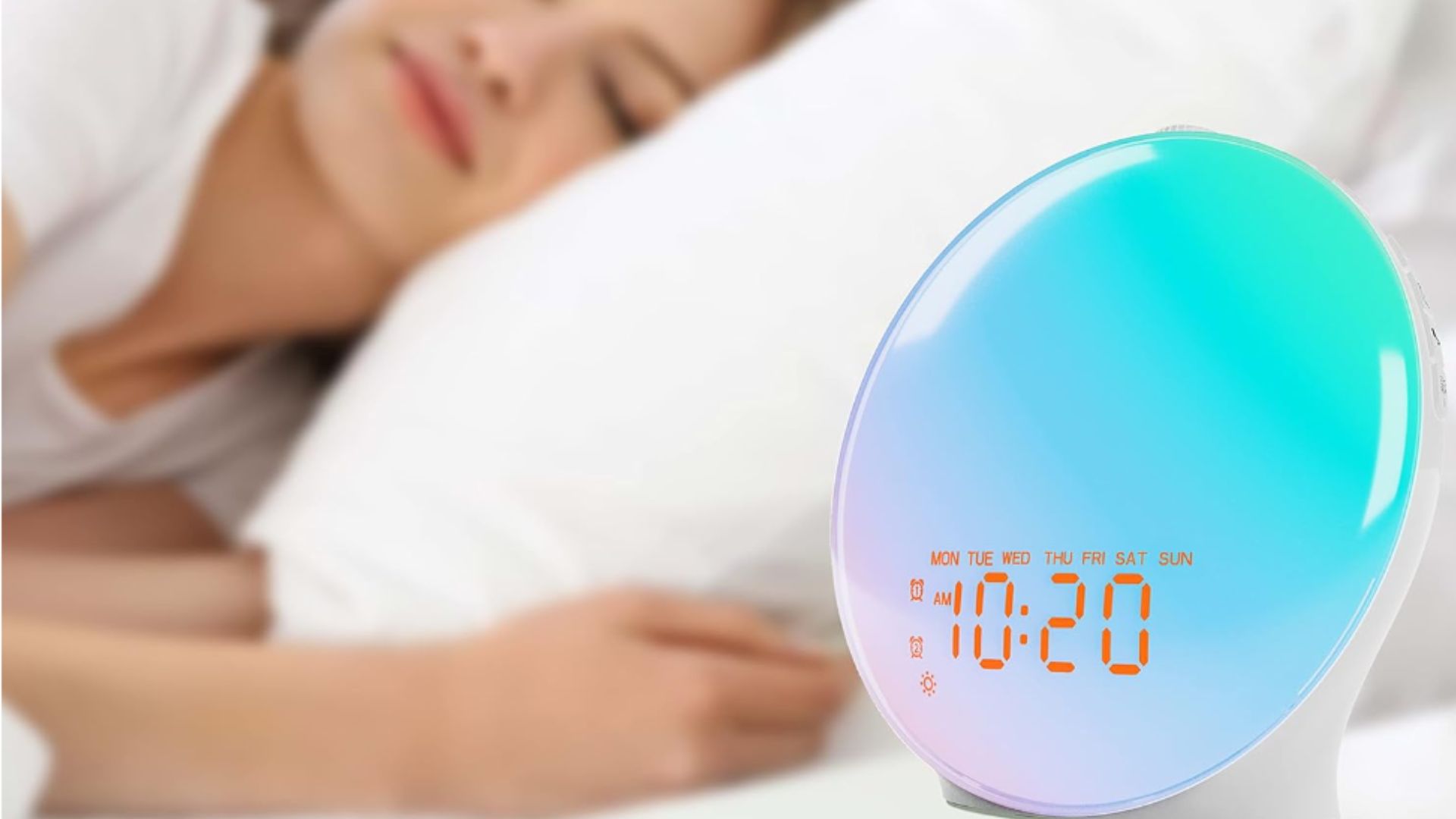 Blue and purple JALL sunrise alarm clock on bedside next to woman in bed sleeping on her side.