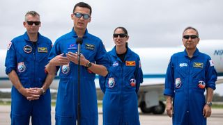 four astronauts in a row with flight suits on. one of them speaks at a microphone while three stand in the background. all are wearing sunglasses