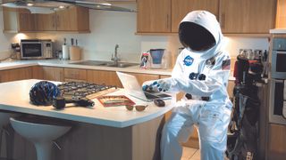 a person dressed as an astronaut sits at a kitchen able surounded by NASA inventions and spin-offs now regularly found in the home.