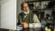 An older man sits at a desk in front of a computer, looking thoughtful.