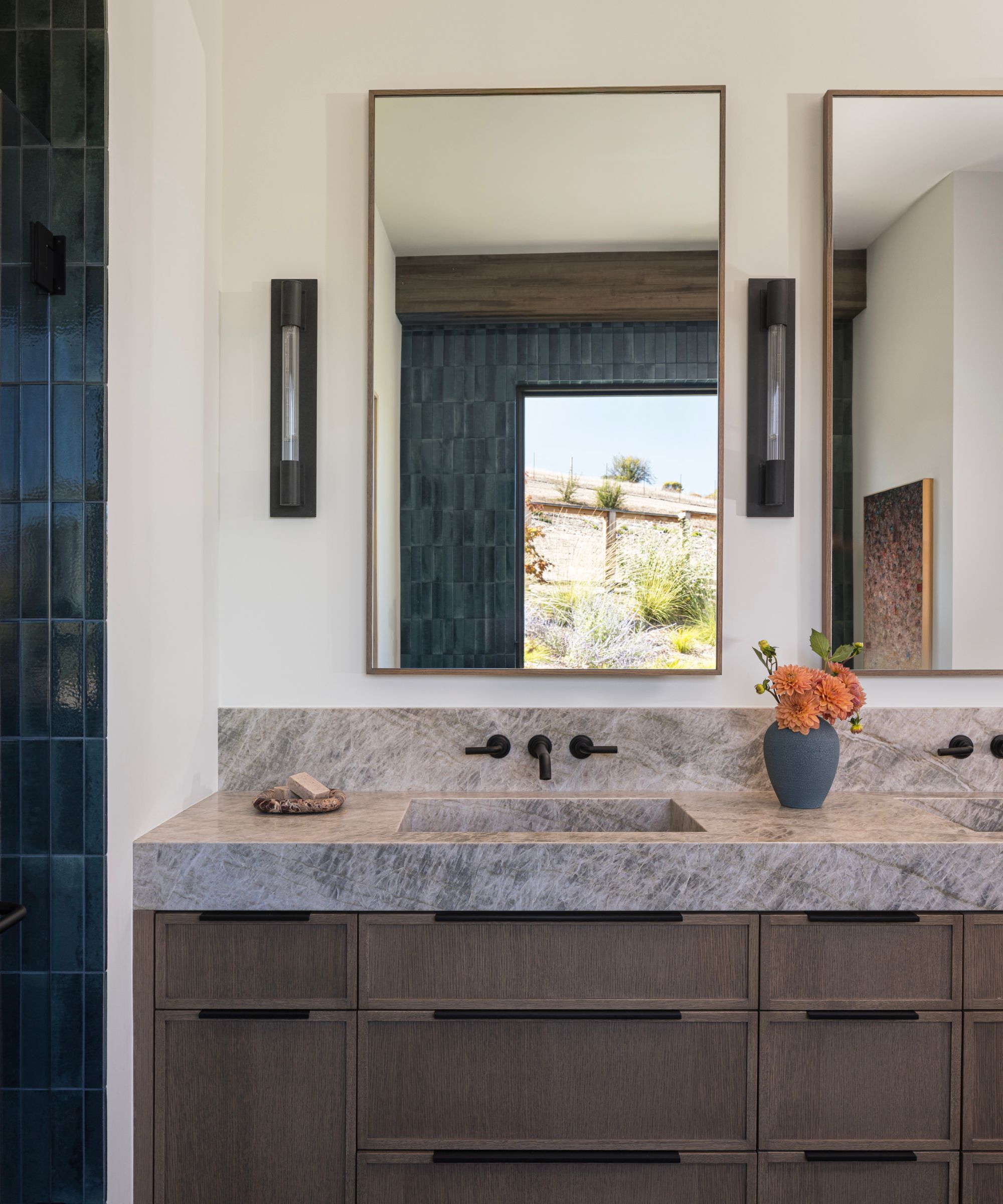 A bathroom with white walls, a wood vanity with a gray marble top, a rectangular brass wall mirror and blue shower tiles