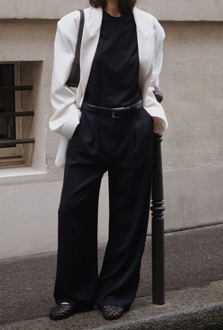 The woven shoe trend is shown in a close street-style photo of a woman standing outside on the sidewalk wearing a boxy white blazer over a black t-shirt with a black belt, oversized black trousers, a black shoulder bag, and black woven ballet flats