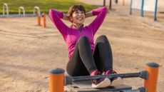 A woman performs assisted sit-ups in a park. Her feet are hooked under a metal pole, her knees are bent and she is sitting up with her hands clasped behind her head. She is smiling broadly; behind her we see a sandy area and grass.