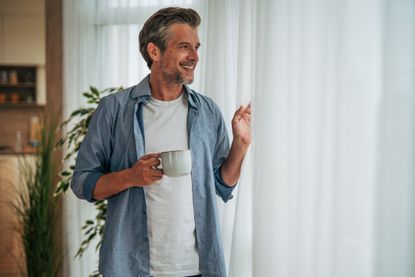 An older man smiles and looks out the window, holding a coffee cup.