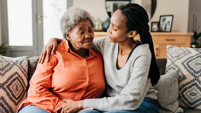 An adult woman hugs her mom while they sit on the sofa together.