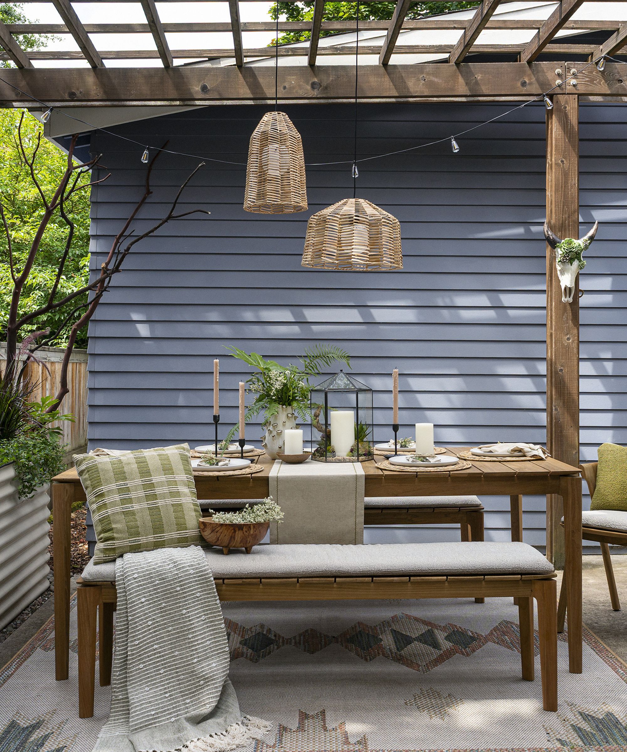 Outdoor wooden table and benches under a pergola with cushions and blankets, overhead rattan lighting and an outdoor rug