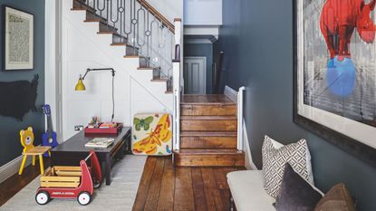 A space with dark blue walls, a white staircase, and dark wood flooring, with a children's playroom area to the left with toys and a low, dark-wood desk on a white rug. To the right is an upholstered bench with mismatched cushions, and a large artwork of a red rhino standing on a blue ball above. The staircase is seen in the background.