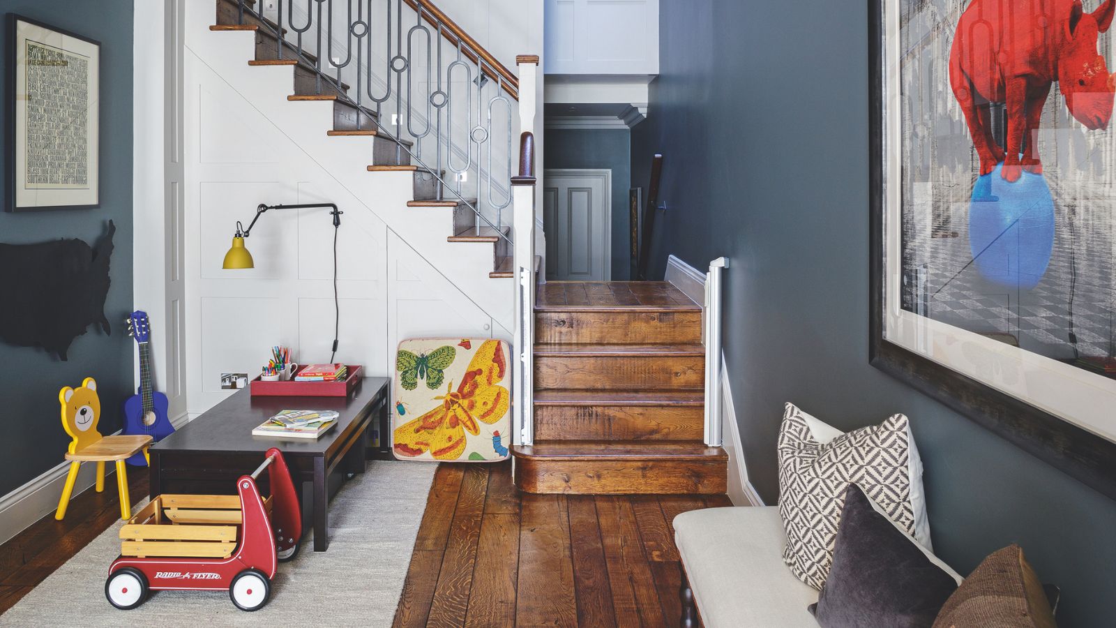 A space with dark blue walls, a white staircase, and dark wood flooring, with a children&#039;s playroom area to the left with toys and a low, dark-wood desk on a white rug. To the right is an upholstered bench with mismatched cushions, and a large artwork of a red rhino standing on a blue ball above. The staircase is seen in the background.