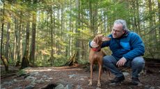 An older man hiking in the woods squats to talk to his dog. 