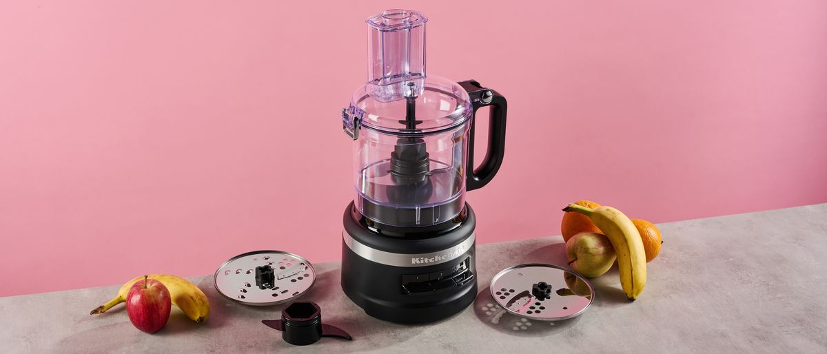 A matte black KitchenAid 7 cup food processor is pictured against a pink background. It is sitting on a grey stone-effect surface on which also sites the reversible slicing and grating disc on the left, with the plastic dough hook positioned just in front, and to the right, the thick slice disc that is only available with the KFP0719 configuration.