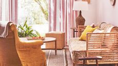 A bright and sunny pink living room with a large window, white and pink striped curtain, rattan couch, mustard armchair, round coffee table, and a rattan lamp with white lampshade.