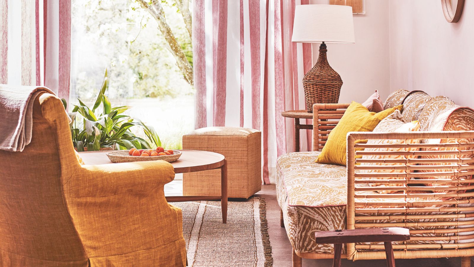 A bright and sunny pink living room with a large window, white and pink striped curtain, rattan couch, mustard armchair, round coffee table, and a rattan lamp with white lampshade.