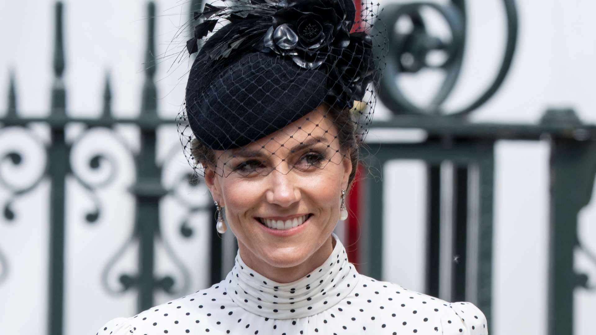 Kate Middleton wearing a white polka dot dress and black hat smiling