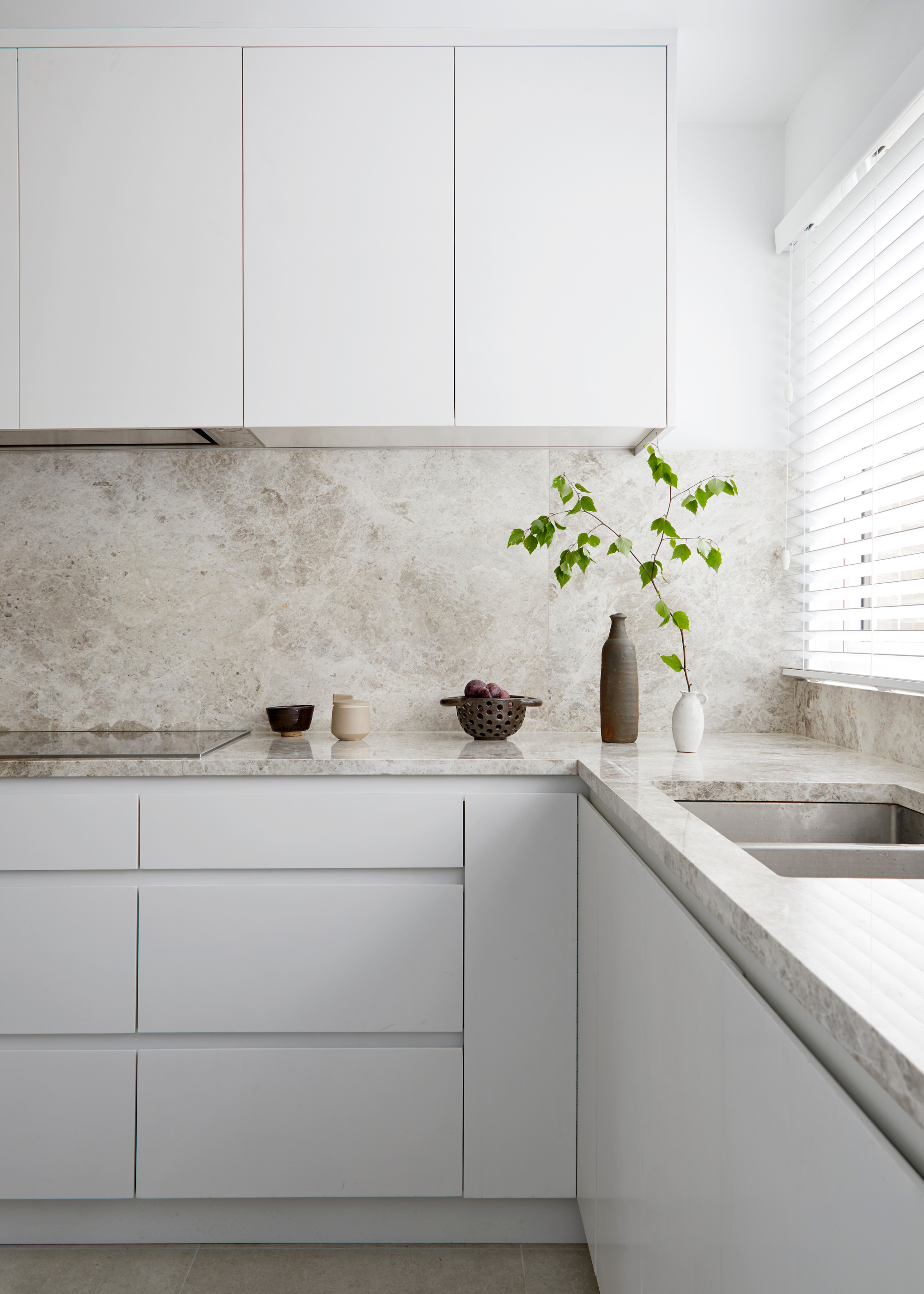 A bright kitchen with white cabinets, a marble counter, a bowl of fruit, a couple of ceramics, and a vase with a stem