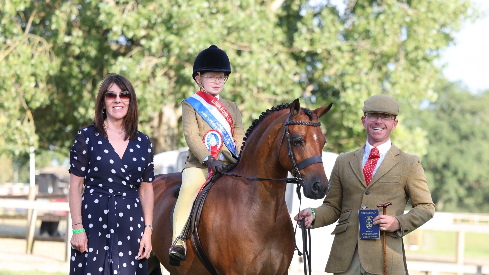 Bay show hunter pony lead-rein Newoak Midnight Blue stands for a photo with his rider Arabella Corrigan on board, held by Adam Forster with a sponsor alongside him.