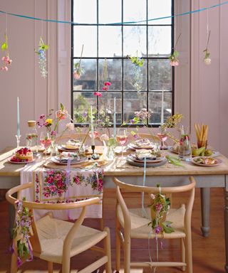 Spring dining area with two wooden curved chairs and a dining table with candles, pastel flatware, wine glasses with pink liquid, and a floral table runner. It has pale pink walls and large black windows.