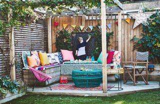 a bright, multi-colored wooden pergola, with multi-colored pillows, garden furniture, bunting, and a large wooden bench against the wooden fences