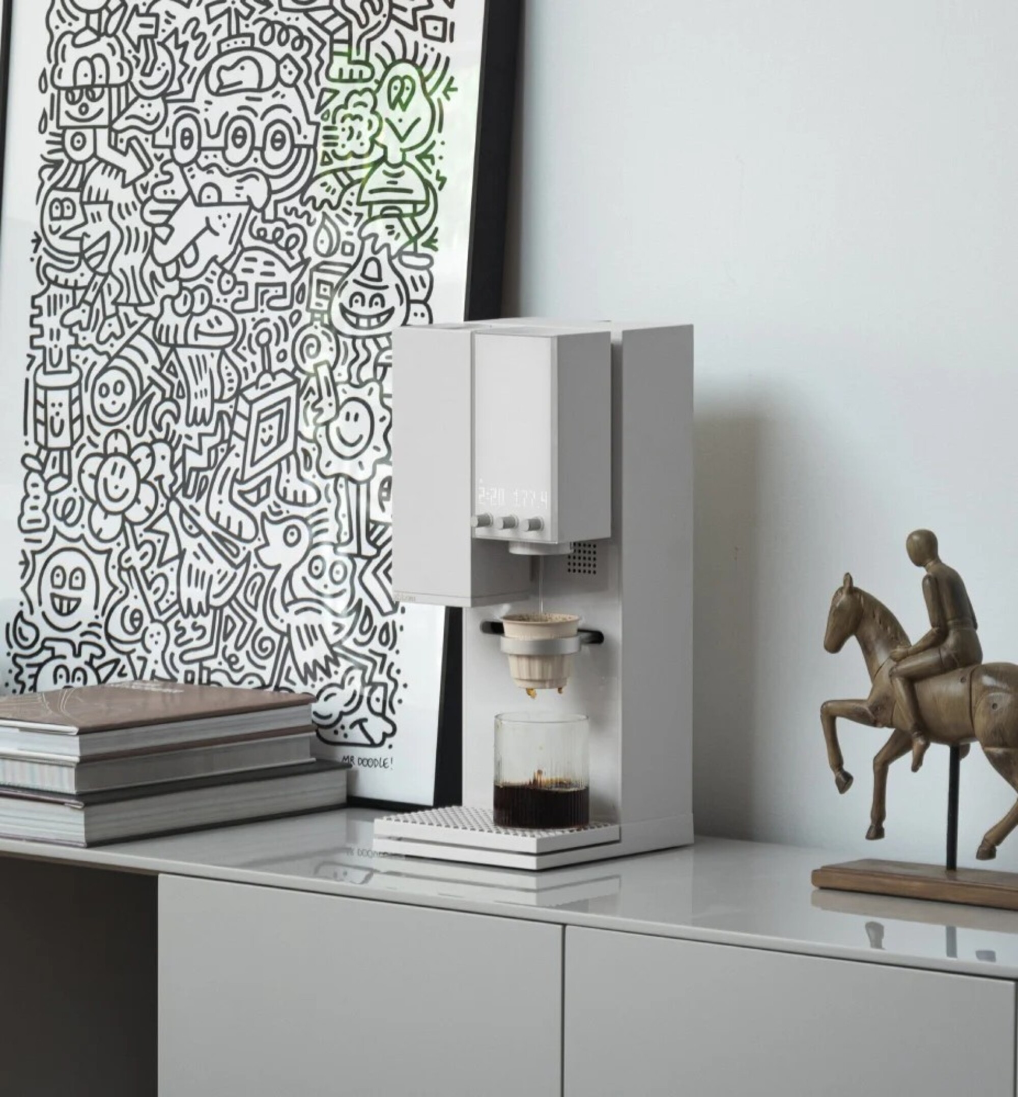 A white pour-over coffee machine by xBloom on a white console in a white room with a black-and-white framed illustration and a horse statue next to it.