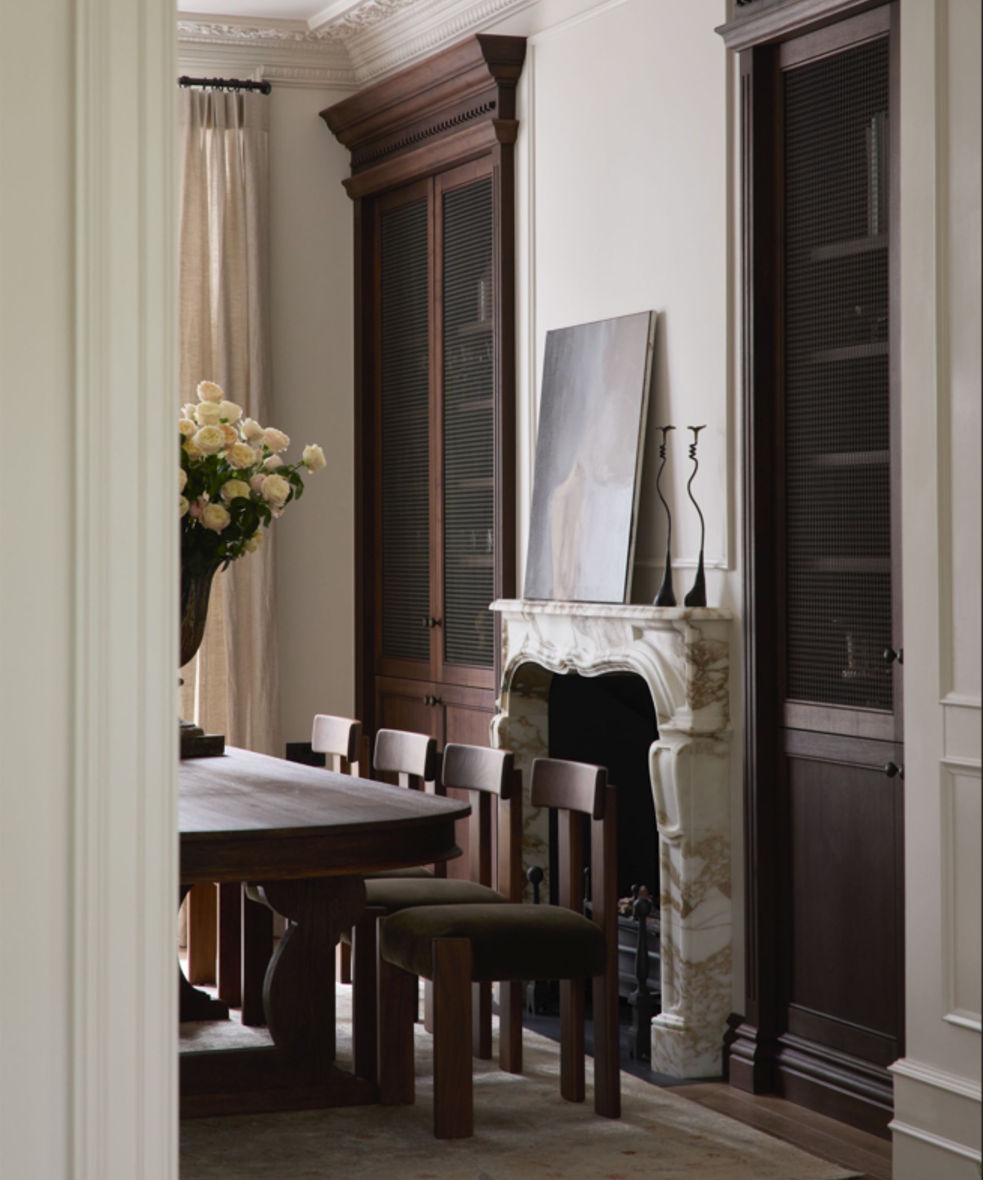 Dining room shot through a doorway, build in wooden alcove cabinetry, marble fireplace with artwork and candlesticks on mantel, dark wooden dining table and chairs