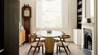 A dining room with a round glass-topped table with floor chairs around it and a pendant light hanging above in front of a fireplace with a grandfather floor clock in the corner and dark wood flooring