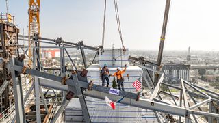 two construction workers pose on steel beams atop a building in a sunny city