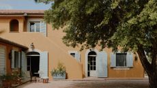 Exterior of a pale orange house with pale blue shutters and a large tree in front