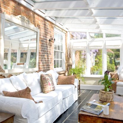 conservatory extension with exposed brick wall, white sofa and armchair, coffee table, grey tiled floor, plants, ochre cushions, large shabby chic style mirror, wall light